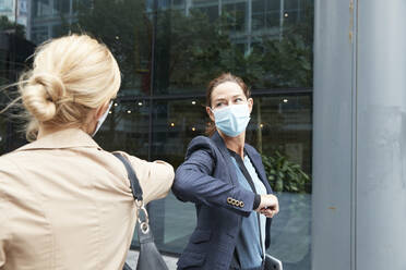 Businesswoman and colleague with face mask greeting with elbow bump while standing against office building - PMF01370