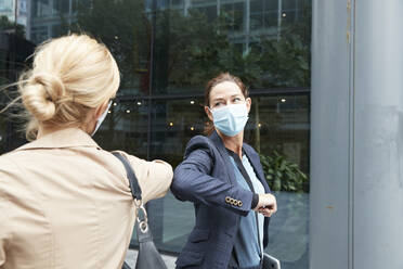 Geschäftsfrau und Kollege mit Gesichtsmaske grüßen mit Ellbogenstoß, während sie vor einem Bürogebäude stehen - PMF01370