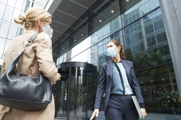 Mature women wearing face mask talking while standing against office building - PMF01371