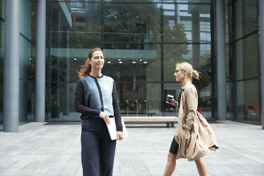 Mature woman standing and holding laptop with colleague walking in background against office building - PMF01381