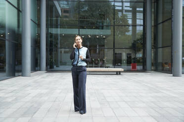 Frau, die mit einem Mobiltelefon spricht, während sie vor einem Bürogebäude steht - PMF01382