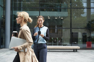 Frau, die einen Laptop hält, während sie mit einer Geschäftsfrau spazieren geht, die im Hintergrund ein Mobiltelefon benutzt - PMF01383