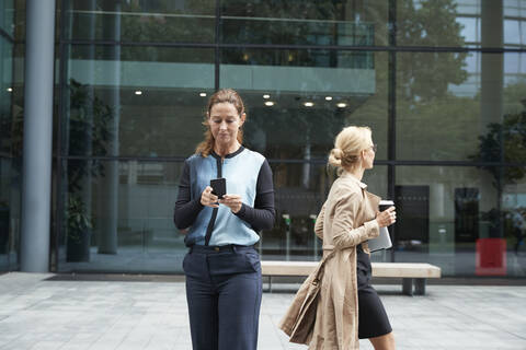 Businesswoman using mobile phone with colleague leaving after work in background stock photo