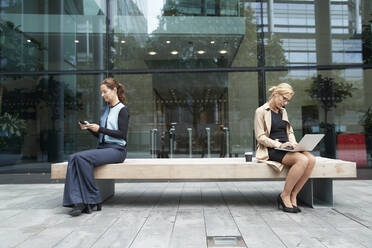Geschäftsfrau und Kollege bei der Arbeit, während sie mit sozialem Abstand auf einer Bank vor einem Bürogebäude sitzen - PMF01385