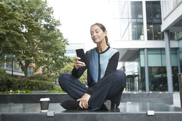 Geschäftsfrau, die ein Mobiltelefon benutzt, während sie auf einer Bank im Büropark sitzt - PMF01386
