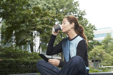 Businesswoman drinking coffee while using mobile phone at office park - PMF01388