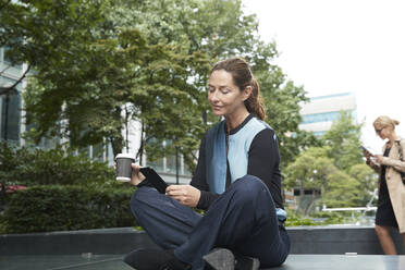 Geschäftsfrau, die eine Kaffeetasse in der Hand hält und ein Mobiltelefon benutzt, während ein Kollege im Hintergrund im Büropark steht - PMF01389