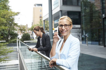 Smiling woman talking on mobile phone while colleague standing in background at city - PMF01394