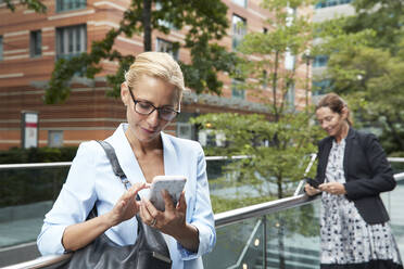 Woman using mobile phone while colleague standing in background at city - PMF01395