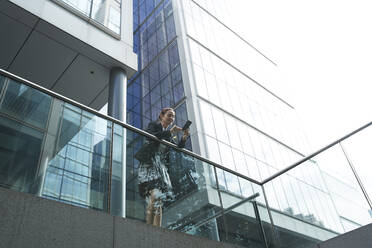 Businesswoman using smart phone while leaning on railing against modern office building - PMF01405