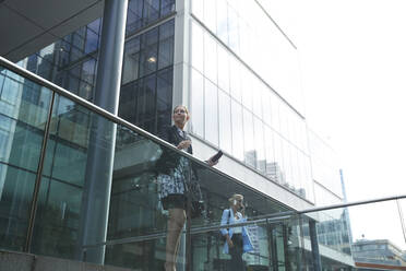 Businesswoman standing by railing with colleague talking on mobile phone in background at city - PMF01409