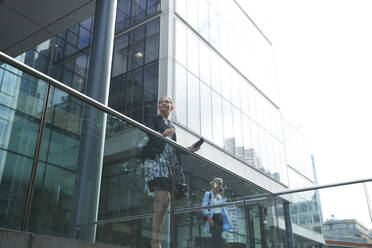 Geschäftsfrau, die an einem Geländer steht, mit einem Kollegen, der im Hintergrund mit einem Mobiltelefon spricht, in einer Stadt - PMF01409