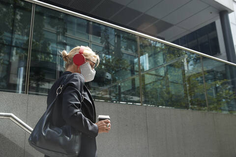 Businesswoman wearing face mask listening music while walking against building stock photo