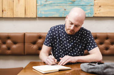 Mittlerer erwachsener Mann, der in einem Café sitzt und in ein Notizbuch schreibt - JMPF00439