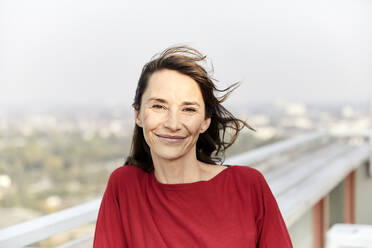 Lächelnde Frau mit wehendem Haar auf einer Gebäudeterrasse stehend - FMKF06478