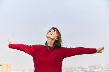 Mature woman looking up with arms outstretched on building terrace against clear sky - FMKF06483