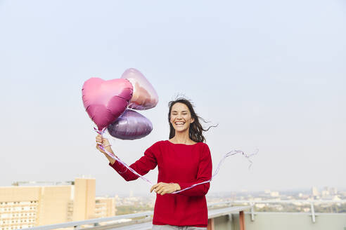 Reife Frau, die einen herzförmigen Ballon hält, während sie auf einer Gebäudeterrasse gegen den klaren Himmel steht - FMKF06489