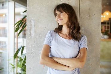 Mature woman with arms crossed standing against concrete wall at home - FMKF06495