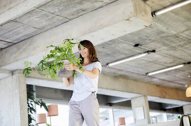 Reife Frau, die eine Topfpflanze trägt, während sie zu Hause steht - FMKF06503