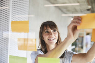 Woman smiling while sticking sticky notes on glass material - FMKF06513