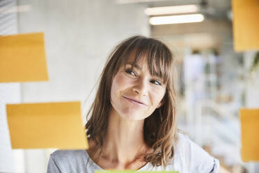 Brown hair mature woman looking at adhesive notes stick on glass material - FMKF06516