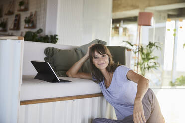 Reife Frau mit Händen im Haar, die ein digitales Tablet benutzt, während sie zu Hause sitzt - FMKF06518