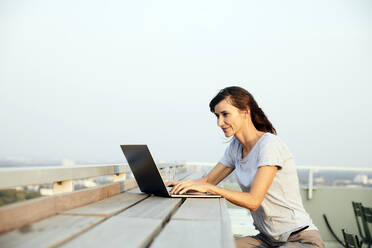 Ältere Frau, die einen Laptop benutzt, während sie auf der Terrasse eines Gebäudes gegen den klaren Himmel sitzt - FMKF06523