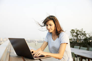 Lächelnde reife Frau, die einen Laptop benutzt, während sie auf der Terrasse eines Gebäudes gegen den klaren Himmel sitzt - FMKF06524