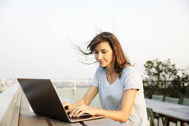 Smiling mature woman using laptop while sitting on building terrace against clear sky - FMKF06524