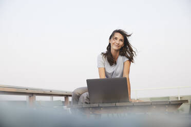 Mature woman looking away while working on laptop sitting on terrace against clear sky - FMKF06525