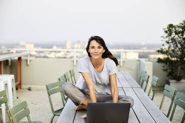 Lächelnde reife Frau im Schneidersitz mit Laptop auf der Terrasse eines Gebäudes sitzend - FMKF06527