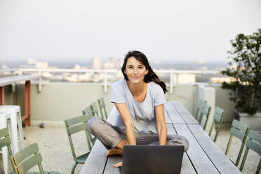 Smiling mature woman with cross legged using laptop while sitting on building terrace - FMKF06527