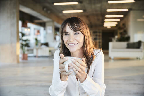 Lächelnde reife Frau hält Kaffeetasse, während sie zu Hause sitzt - FMKF06534
