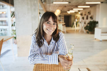 Lächelnde reife Frau hält Bierflasche, während sie zu Hause sitzt - FMKF06540