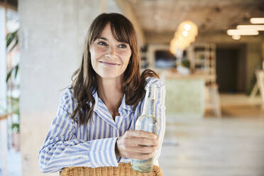 Smiling mature woman looking away while holding beer bottle sitting at home - FMKF06543