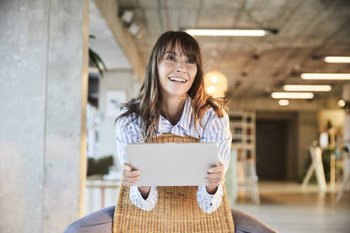 Lächelnde reife Frau, die wegschaut, während sie ein digitales Tablet benutzt, das auf einem Stuhl zu Hause sitzt - FMKF06545