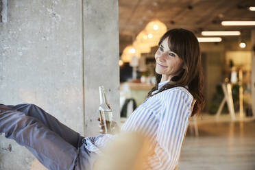 Smiling woman looking away while holding beer bottle sitting on chair at home - FMKF06551