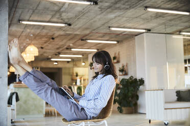 Mature woman listening to music while using digital laptop sitting on chair at home - FMKF06554