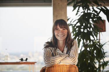 Smiling mature woman with arms crossed sitting on chair at home - FMKF06555