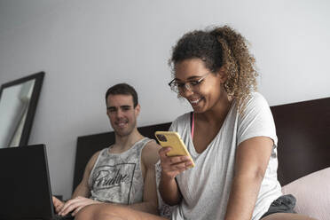 Man working on laptop while looking at woman using mobile phone at home - SNF00620