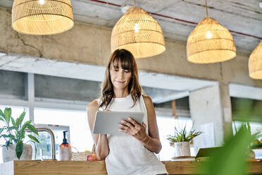 Woman working on digital tablet while standing at home - FMKF06586