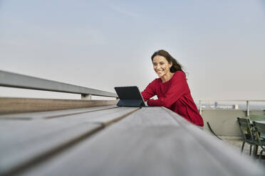 Smiling businesswoman holding digital tablet while leaning on retaining wall at rooftop - FMKF06590