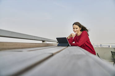 Lächelnde Geschäftsfrau, die ein digitales Tablet in der Hand hält, während sie sich auf eine Stützmauer auf dem Dach stützt - FMKF06591