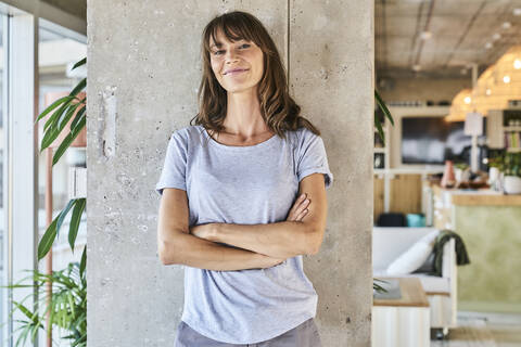 Confident woman standing with arms crossed against wall at home stock photo