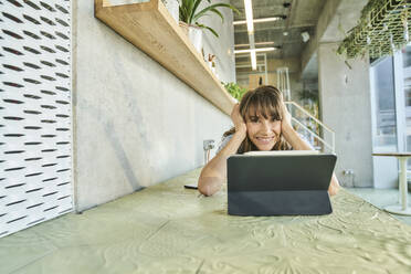 Smiling woman with head in hands using digital tablet while lying on table at home - FMKF06623