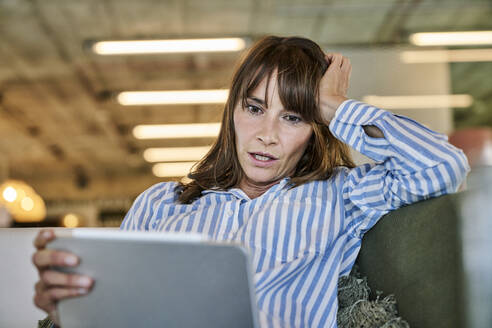 Frau mit schockiertem Gesichtsausdruck, die auf ein digitales Tablet schaut, während sie zu Hause auf dem Sofa sitzt - FMKF06629