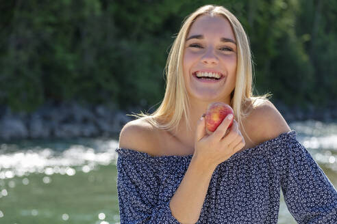 Young woman with apple laughing while standing by river at forest - LBF03238