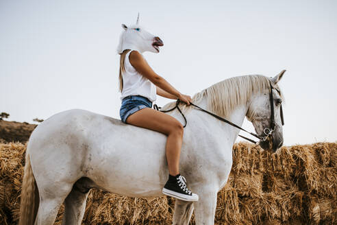 Young woman wearing unicorn mask sitting on horseback - MIMFF00252