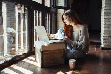 Lächelnde rothaarige Frau, die in ein Buch schreibt, während sie zu Hause einen Laptop benutzt - JSMF01810
