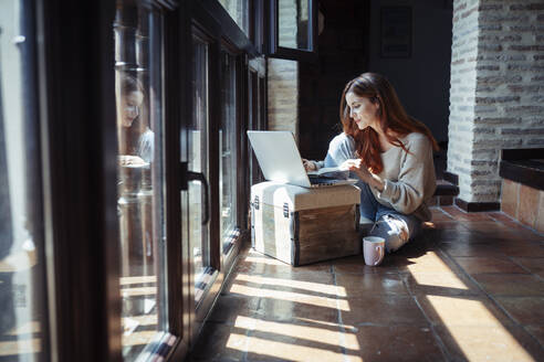 Junge Frau arbeitet am Laptop, während sie zu Hause auf dem Boden sitzt - JSMF01812